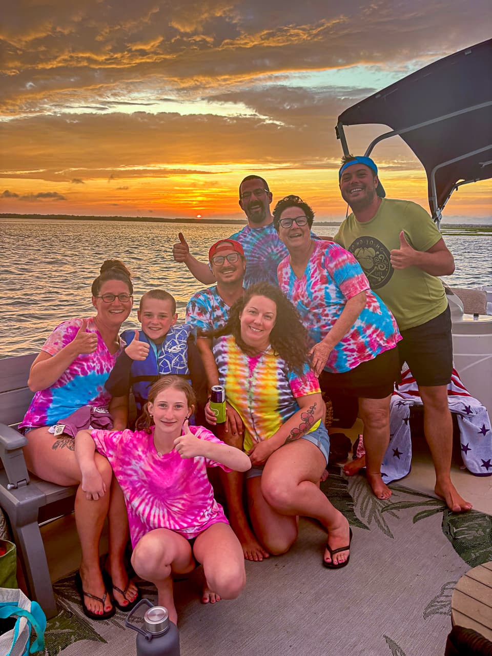 A family in matching tie-dye giving a thumbs up at sunset on a family-friendly Sand Bar Joe's pontoon cruise